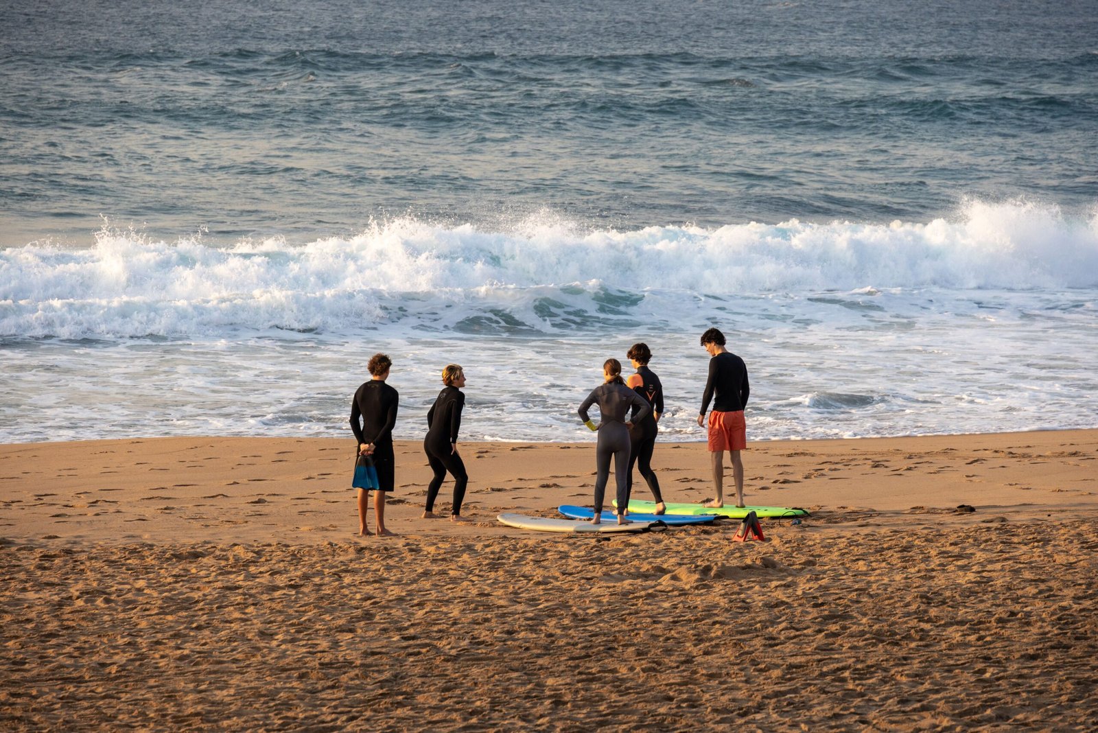 Gallery A group of adults preparing for a surf lesson on a sunny beach with waves crashing in the background.